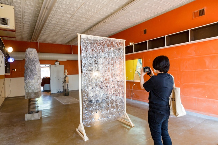 Seulki takes a photo of her installation in the gallery. A large frame holds up multiple small rocks and there are two pedestals holding up two large rock-like objects
