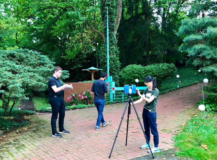 Three students on a brick-paved walkway surrounded by trees, setting up survey equipment.