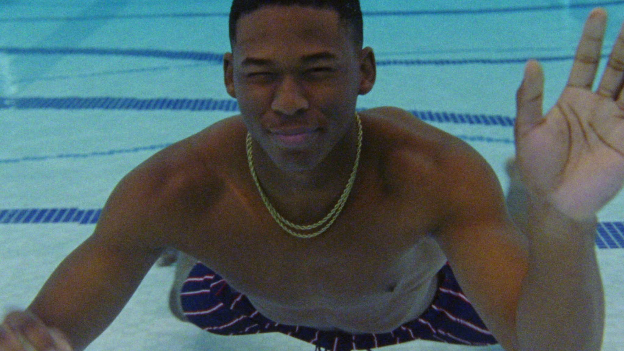 A young man underwater in a pool with one hand up waving