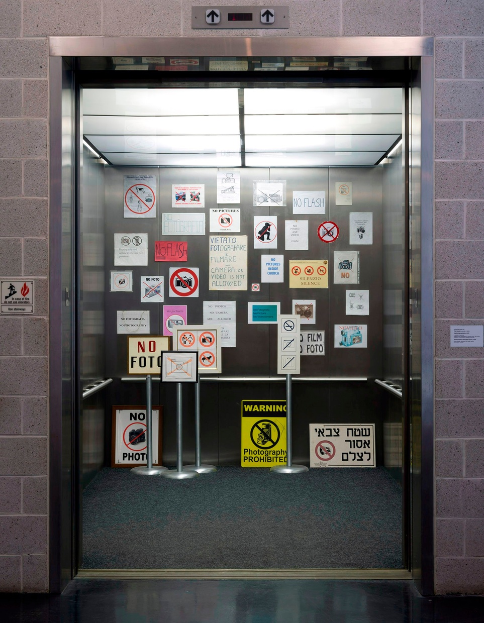 The inside of a metal elevator with numerous posters banning photography in different languages scattered along the walls.