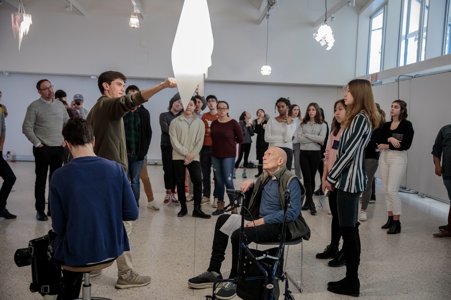 People gathered in a gallery space listen as a student talks about a large pendant light hung from the ceiling.