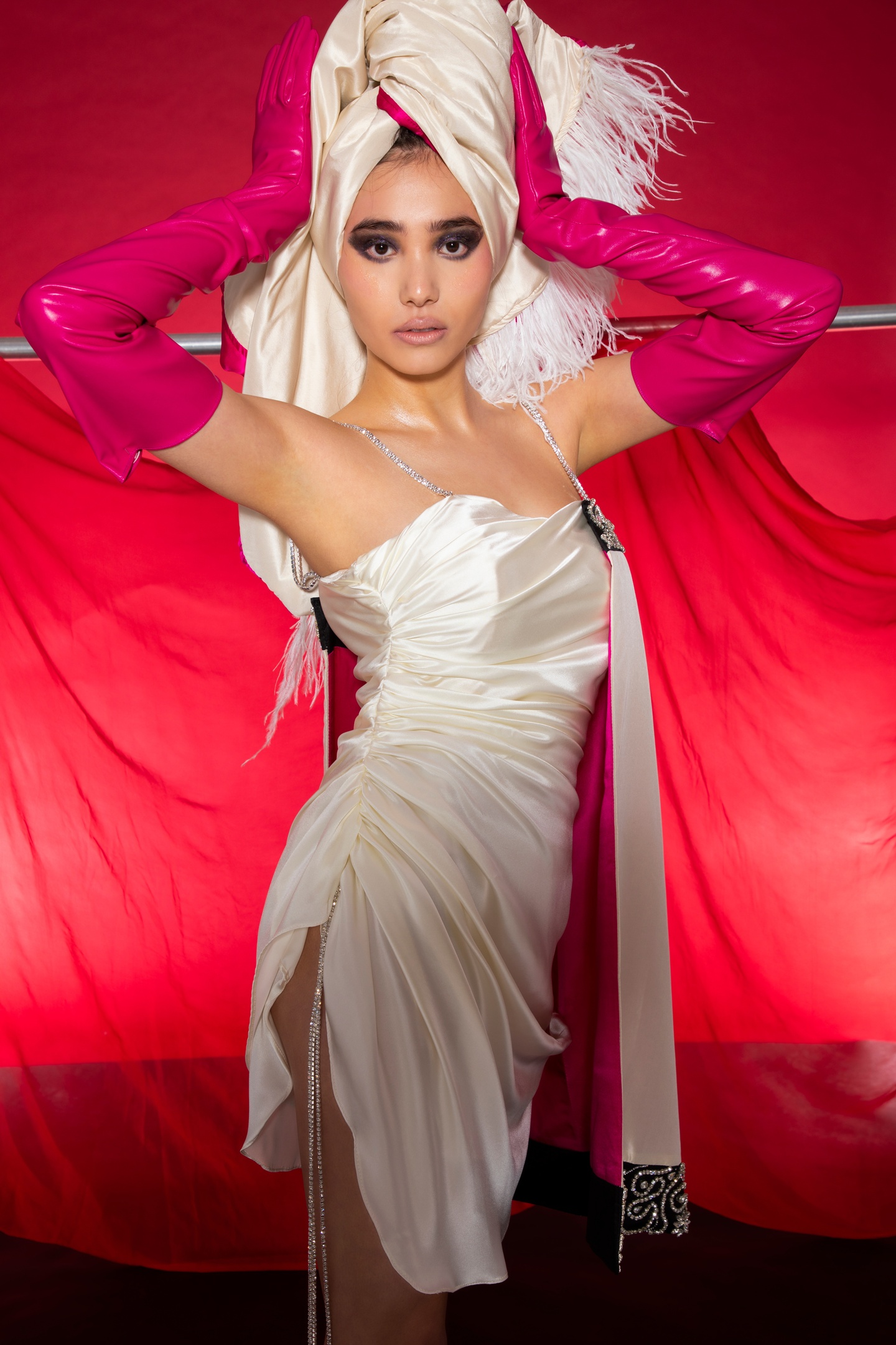 Model wears a short white satin dress cinched up along the side edges to create wrinkles. The dress has two rhinestone studded spaghetti straps and a cape-like panel in white satin lined with magenta attached to the upper left breast. Model wears pink pleather gloves extending all the way up to their biceps and clutches a huge white satin turban with a white feathered fringe on their head.