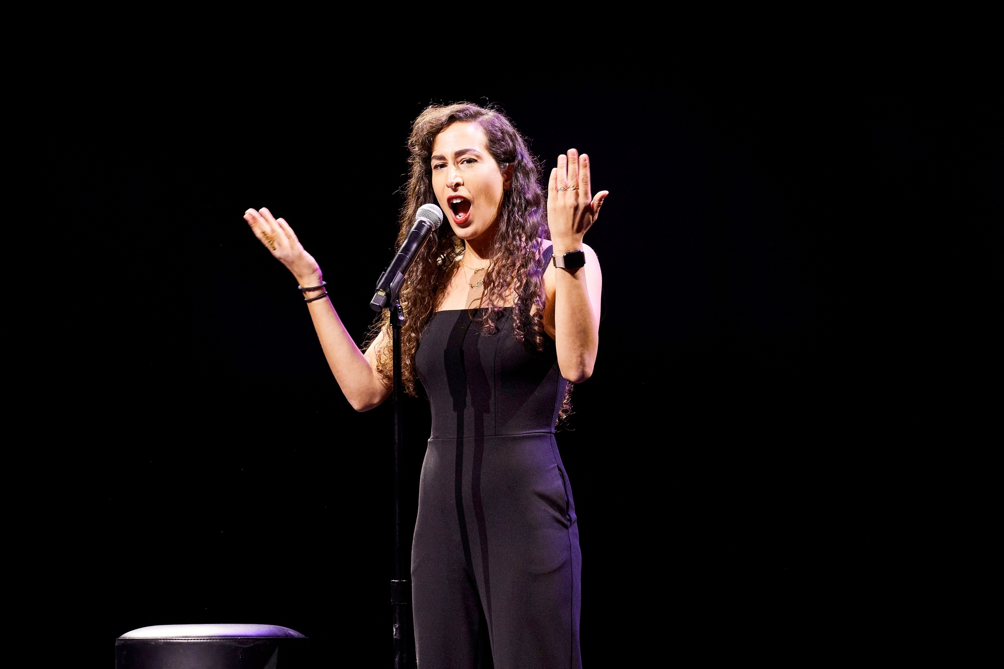 A woman with long brown hair in loose curls holds her hands up in front of her while speaking into a microphone.