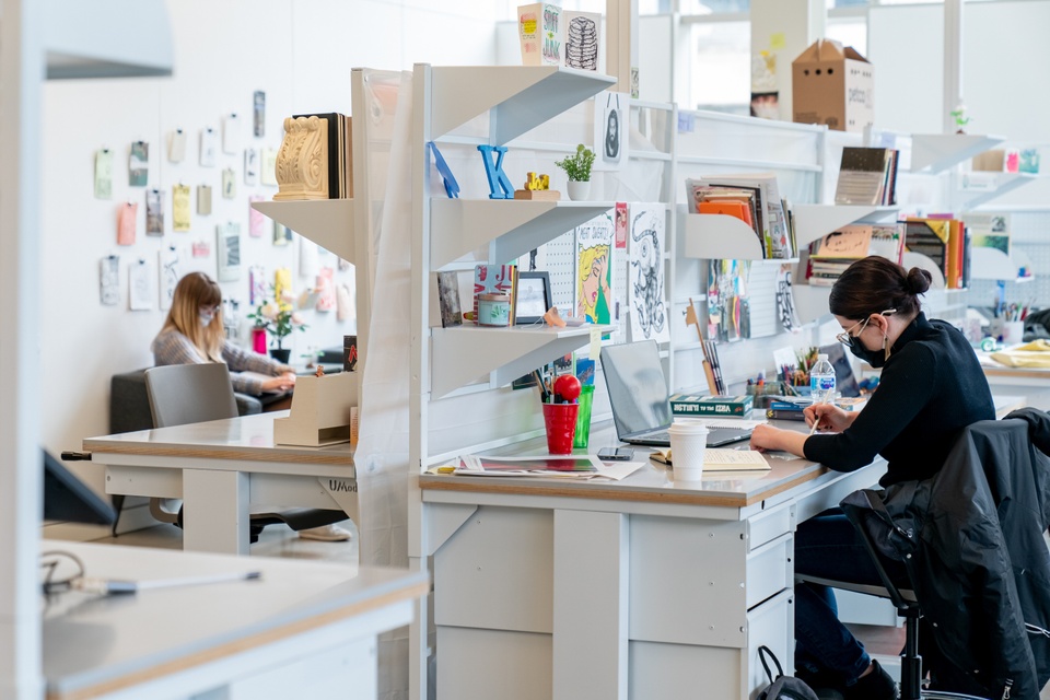 Wide angle shot of an open plan studio space. One person works at their desk, while another sits in an armchair and sketches. Mini comics and zines are pinned up on the wall behind the armchair.