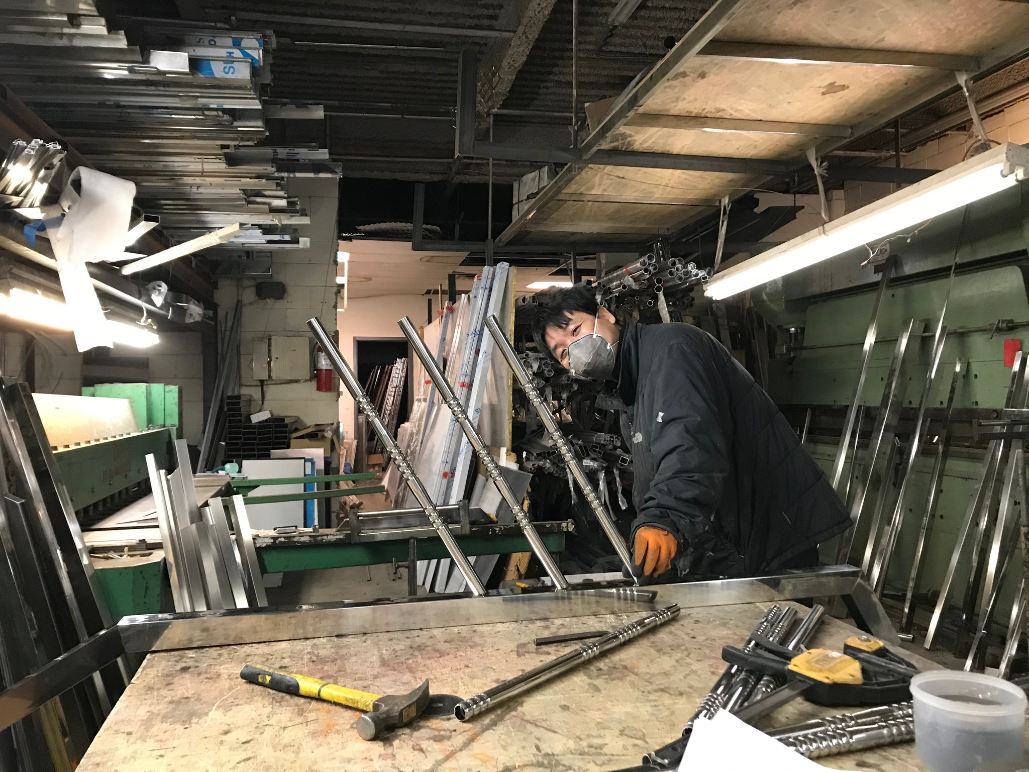 A Chinese man in a cluttered metalworking workshop turns his face toward the camera as he bends over stainless steel railings and tools in front of him. He wears a protective face mask over his mouth and nose and orange gloves on his hands, one of which is stretched out in front of him.   