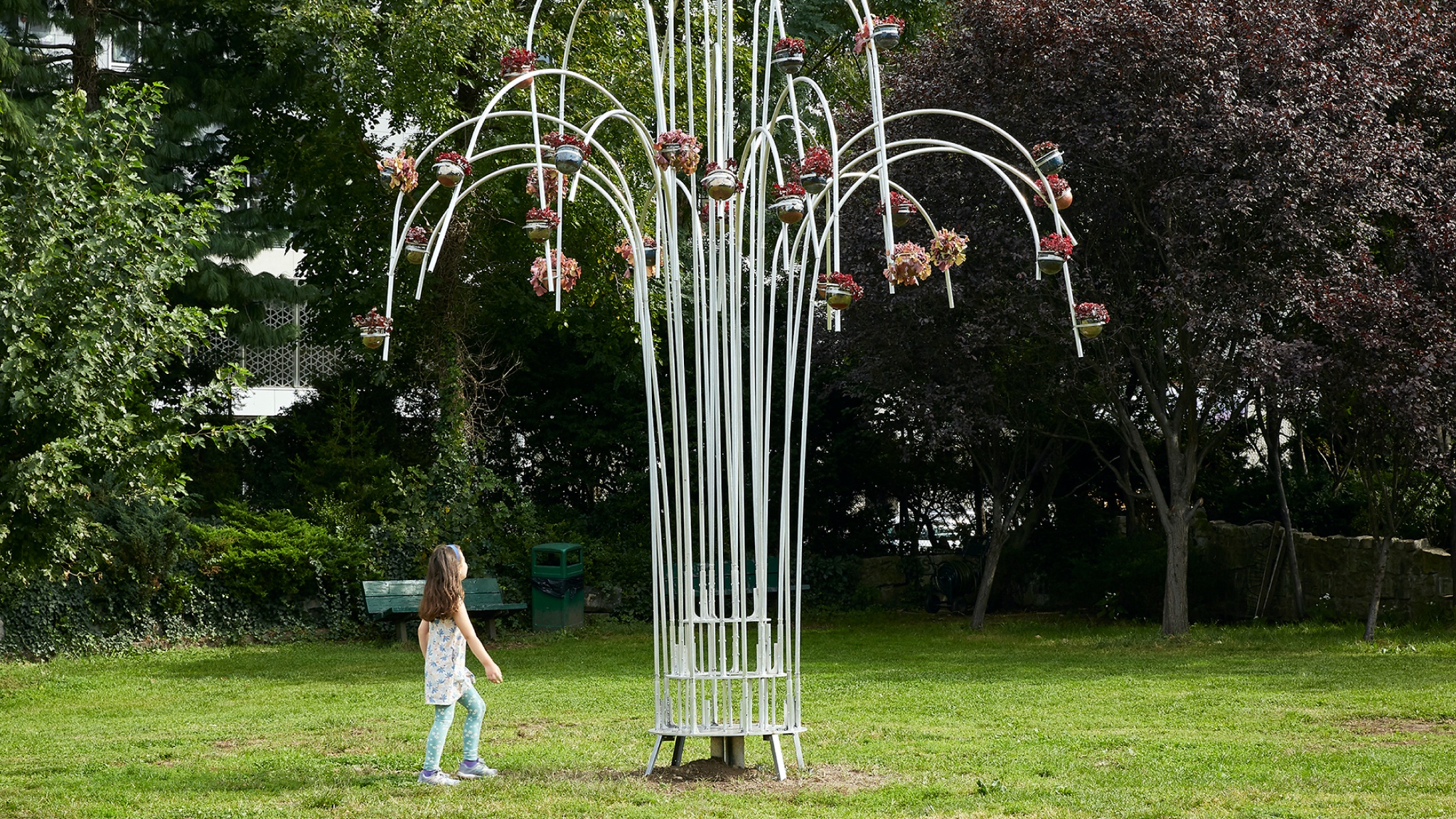 A little girl approaches a sculpture in the middle of a grassy lawn. The sculpture is made of aluminum pipes the stretch upward vertically before bending over slightly at their ends, in the shape of water shooting from a fountain. At the ends of the pipes are coral bell plants held in suspended clayware pots. 