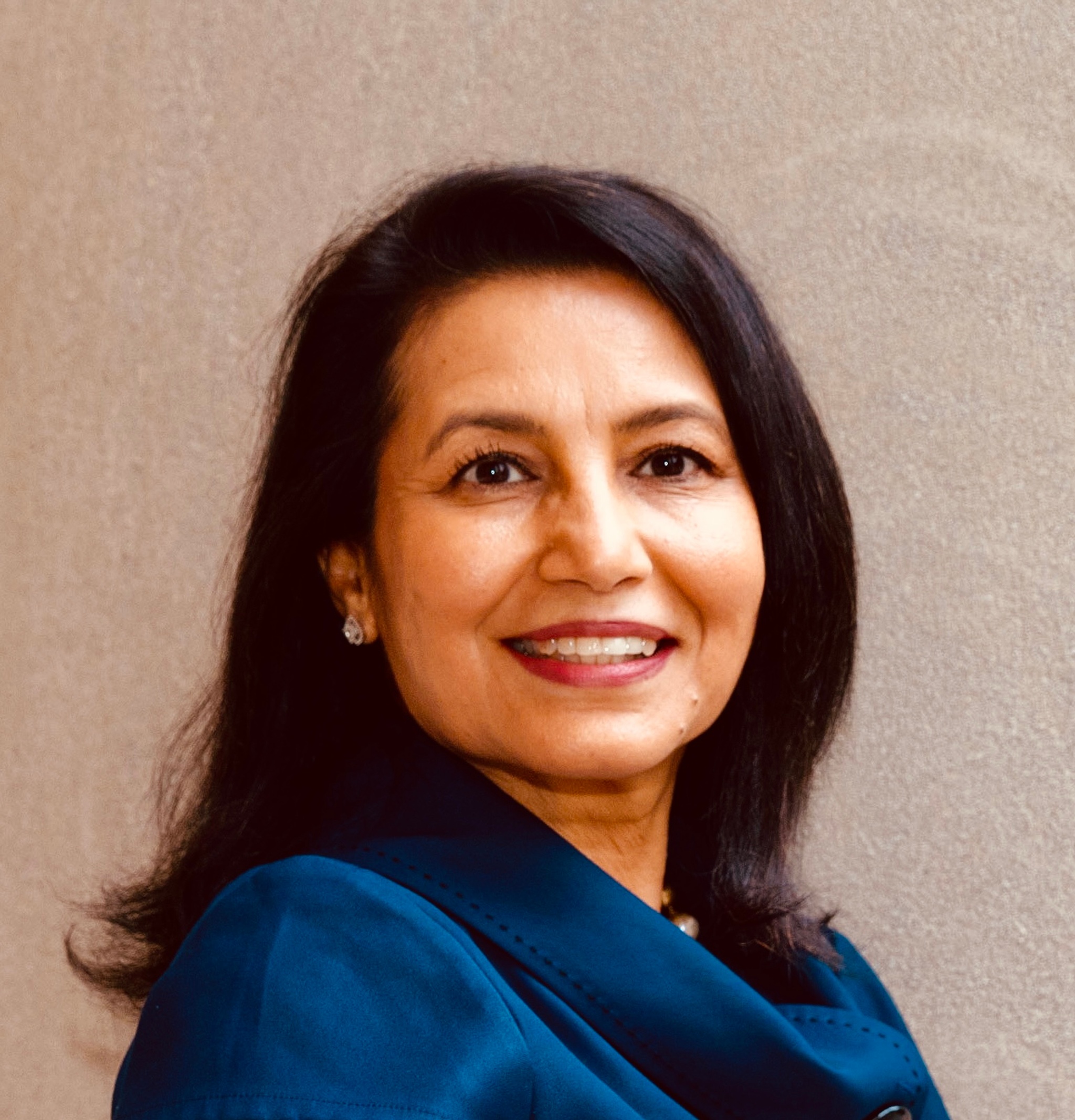 A headshot of Purnima Kapur, who had shoulder-length brown hair, and turns her head to look at us while smiling. She wears a royal blue blouse with short sleeves and a cowl neck.