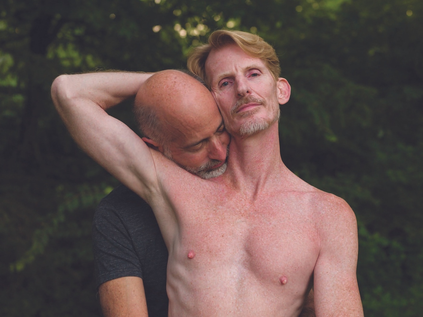 A shirtless person shown from the chest up, standing in front of another person with their arm cradling the head of the person standing behind.