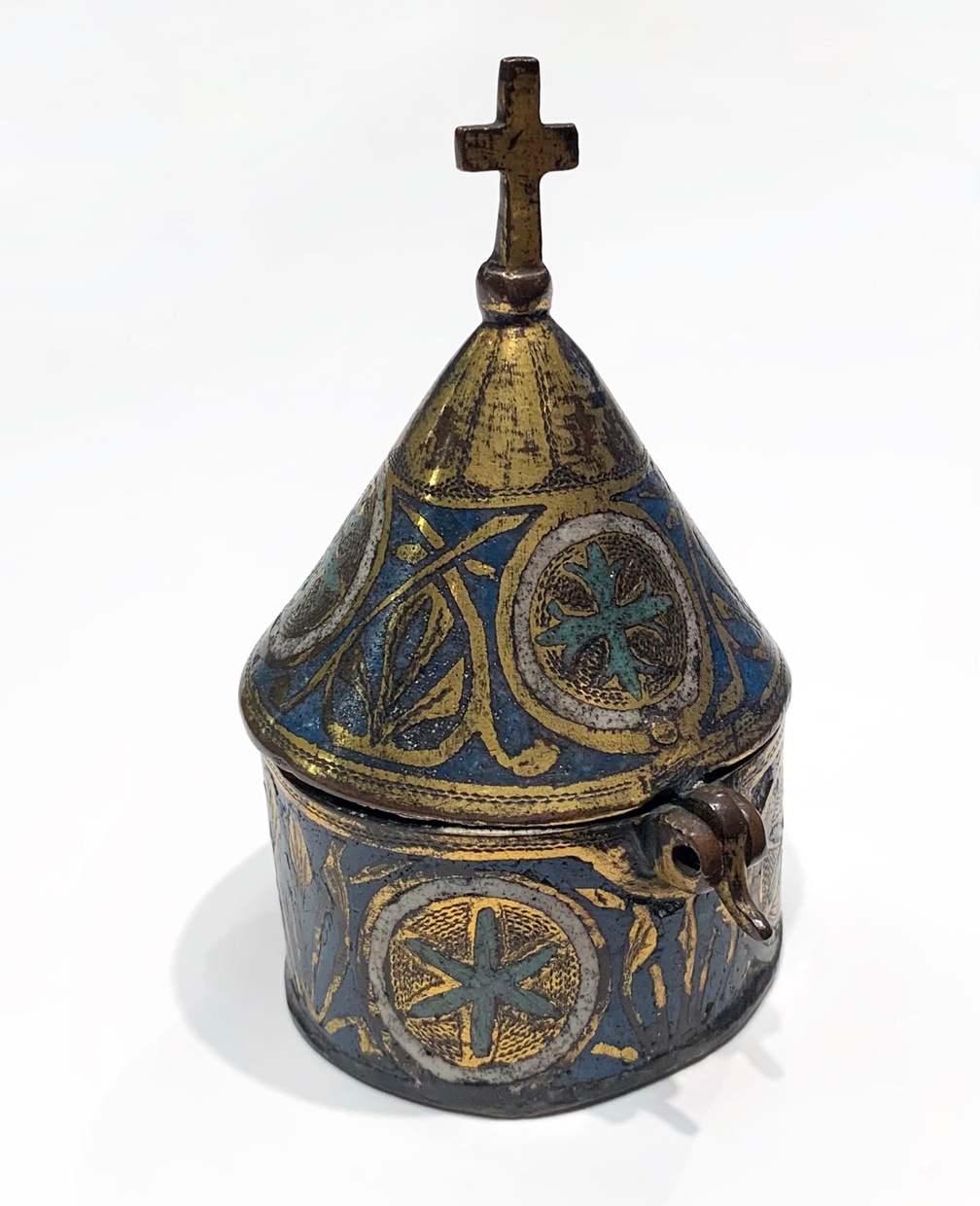 A small round container with blue and gold patterns and a gold cross at the top of a funnel-shaped lid.
