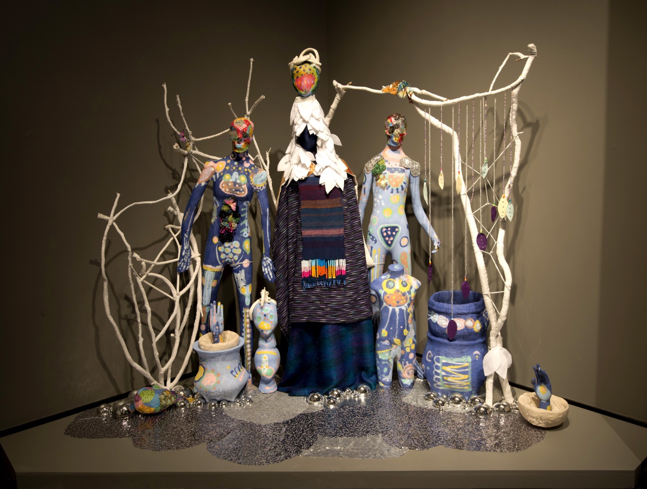 Two, abstractly painted, humanoid sculptures flank another humanoid sculpture wearing multiple, woven blankets and a hood of white petals surrounded by decorative clay vessels and woven tree branches.
