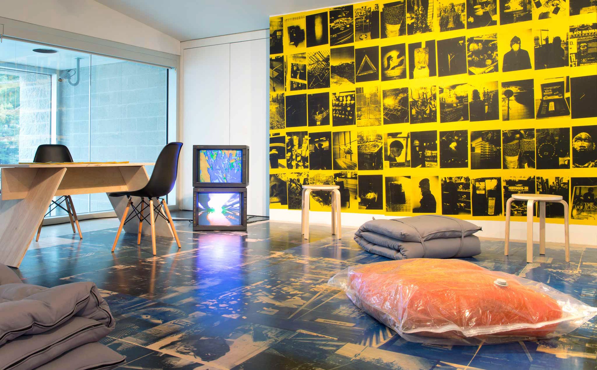 Two TV monitors and various types of seating rest on a wooden floor printed with dark images. Adjacent to the TV monitors, a wall is covered with yellow pamphlets printed with different urban scenes printed in black ink.