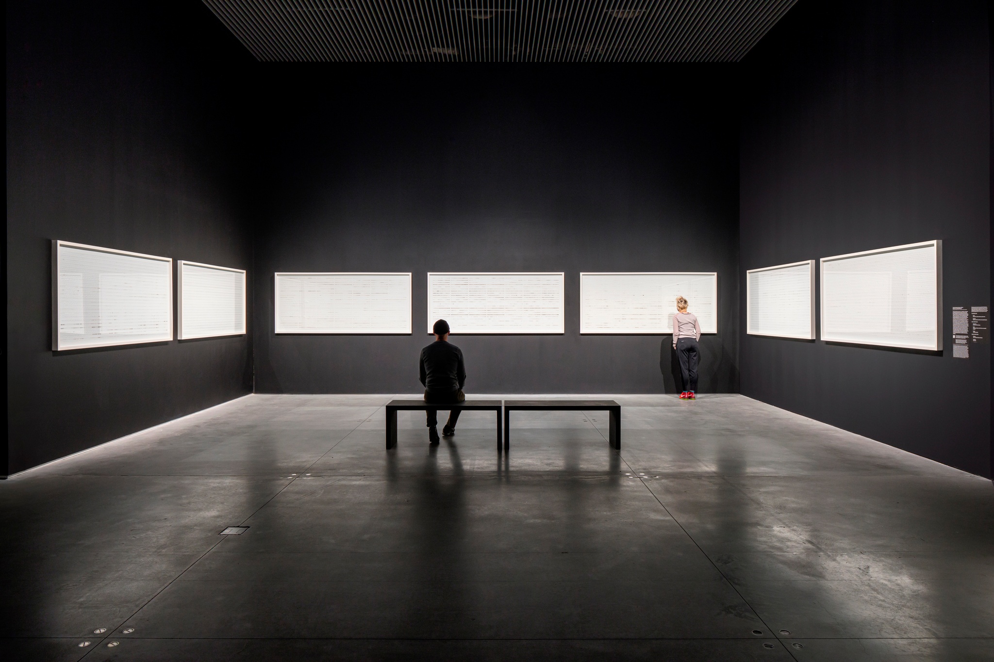 Seven large horizontal frames line three walls of a gallery space. A person sits on a bench centered in front of them and another person stands close to one frame on the left, examining its contents.