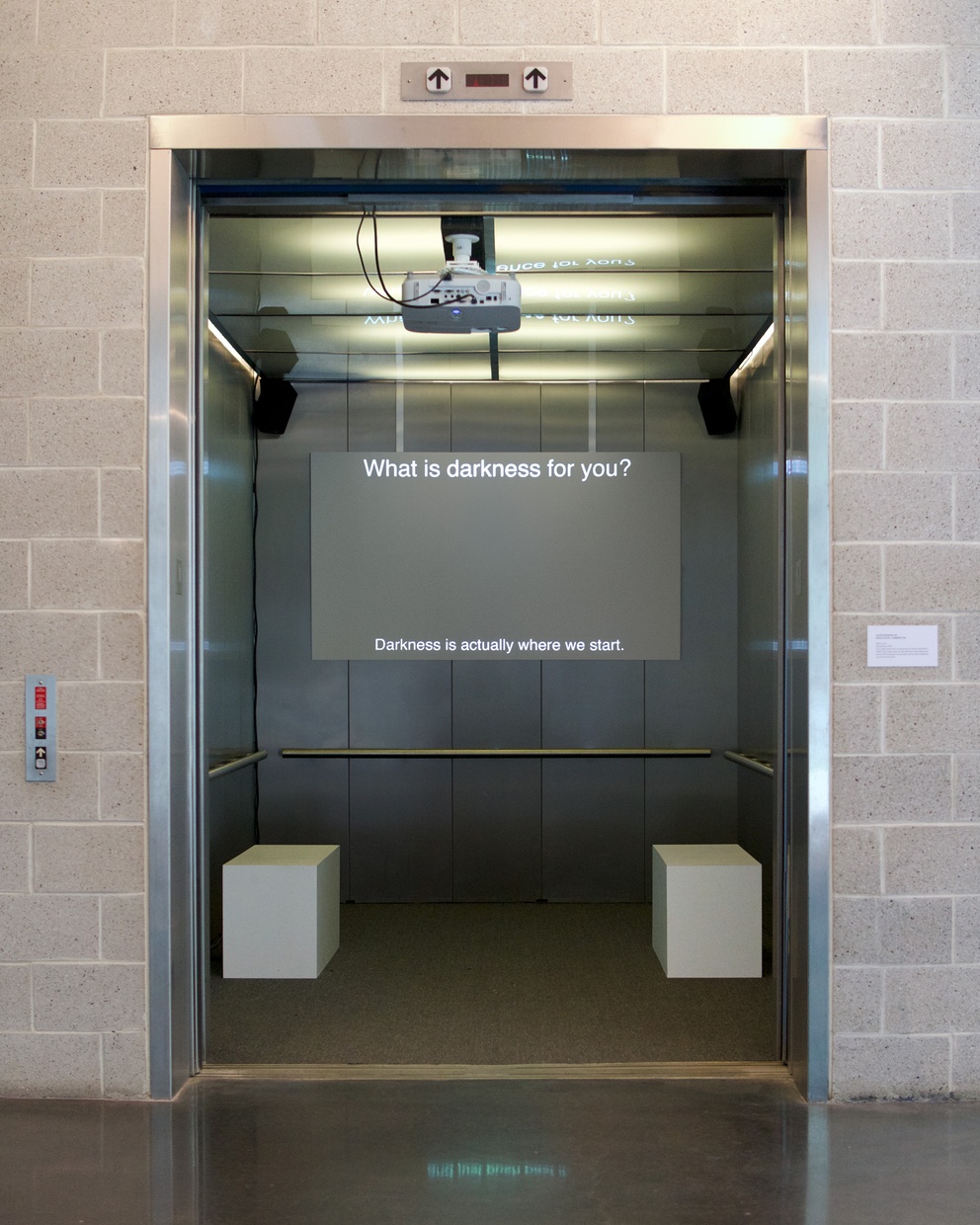 On the back wall of an open elevator a video still is projected on a screen and two white stools are placed parallel to each other along the side walls.