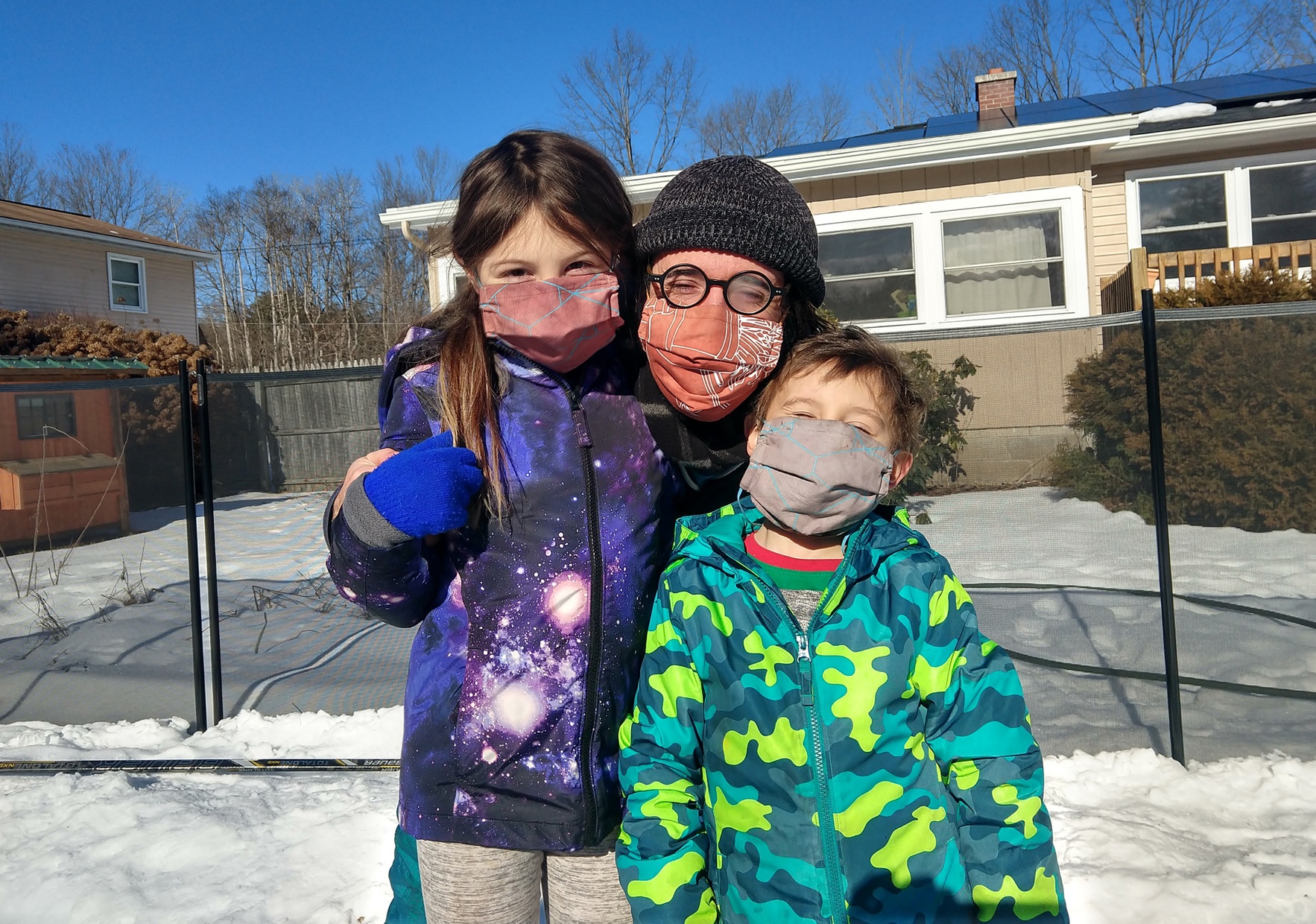 A light-skinned man wearing round glasses and a red mask poses in between two, young light-skinned children wearing snow clothes also wearing masks.