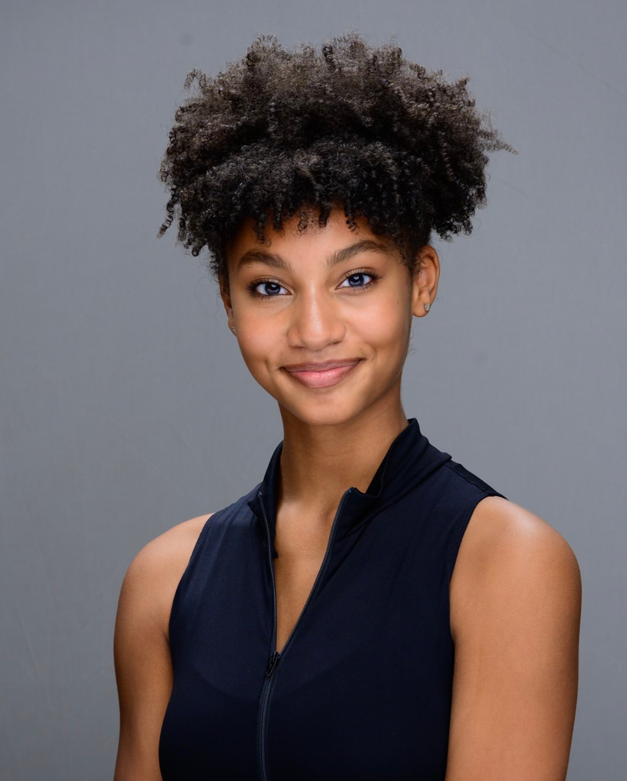 A portrait of dancer Amya Fortunato, a Black person who has curly dark hair pulled back above her head. She has light brown skin and smiles with eyebrows raised at us. She wears a blue sleeveless blouse with a high collar. 