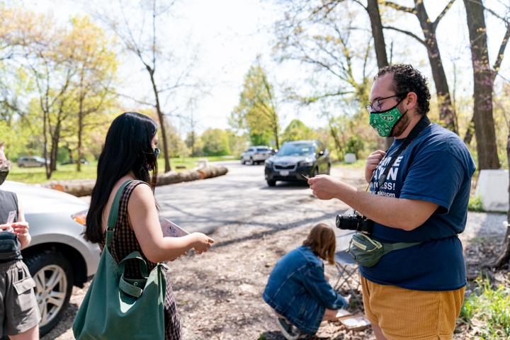 Two people in facemasks in a park. One is handing the other a manilla tag.