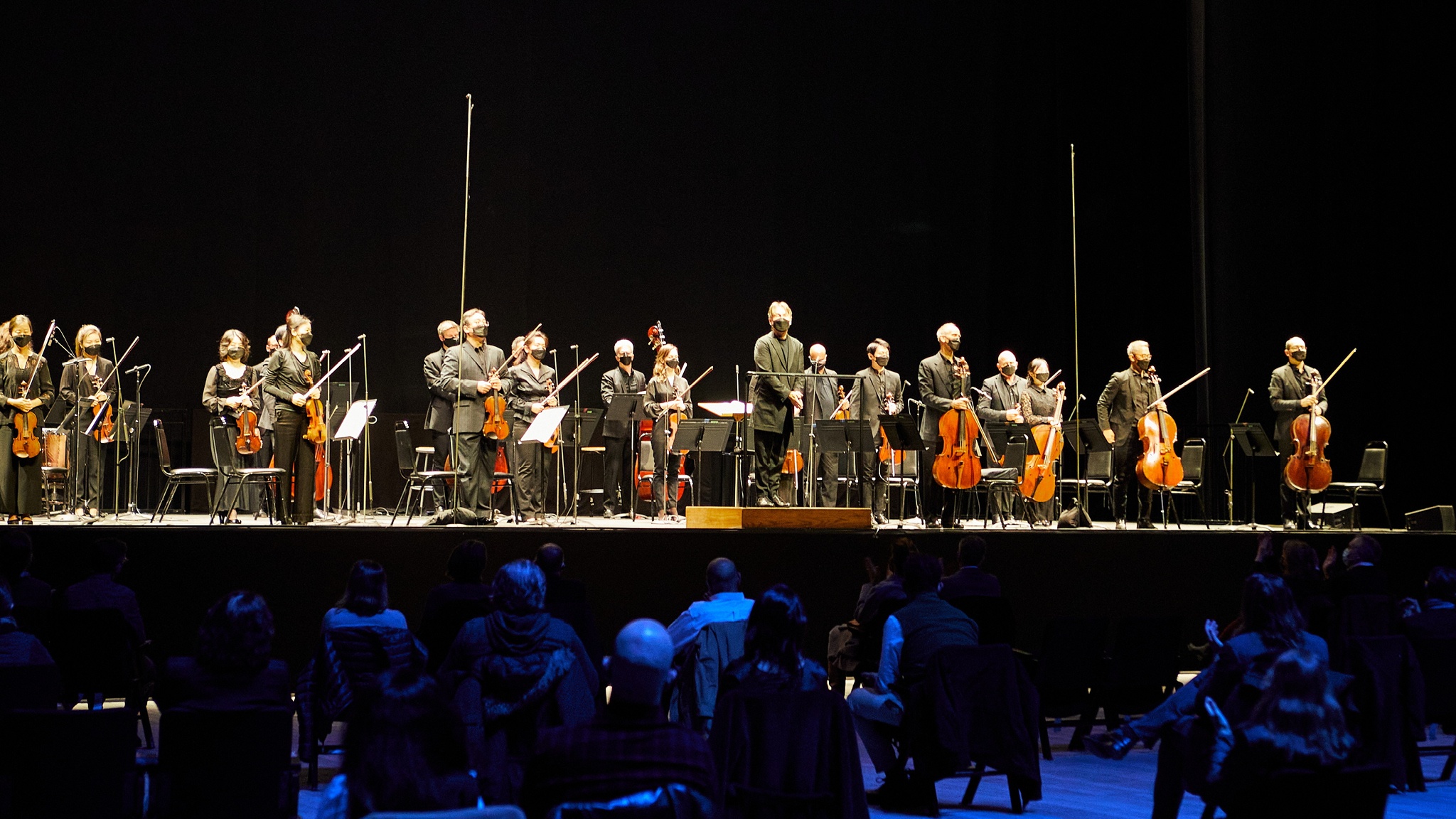 An orchestra standing on a raised stage with people sitting on the floor in the audience in front of them