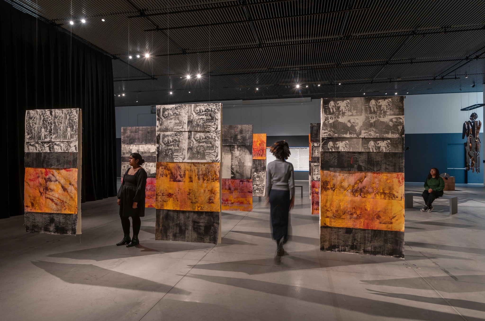 Nine painted fabric panels hang from a gallery ceiling in a grid. Two gallery visitors walk between the panels. The panels are painted in orange and black bands, with screen printed sections with black and white images of banana plantation workers in the 1920s.