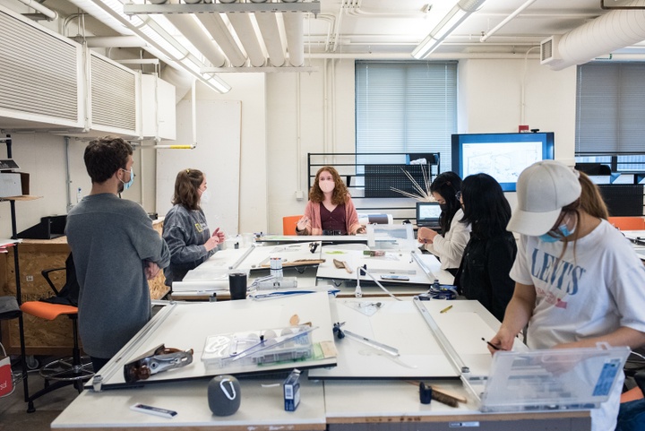 Open studio space with long work tables covered with drafting boards. Several students sit and chat, some work on drawings.