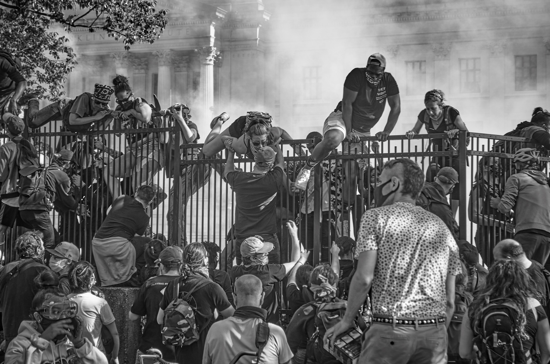 A black and white photograph of protestors climbing over an iron fence in front of a colonial-stye building towards a group of protestors on the other side. A white fog curls up behind the protestors climbing the fence.