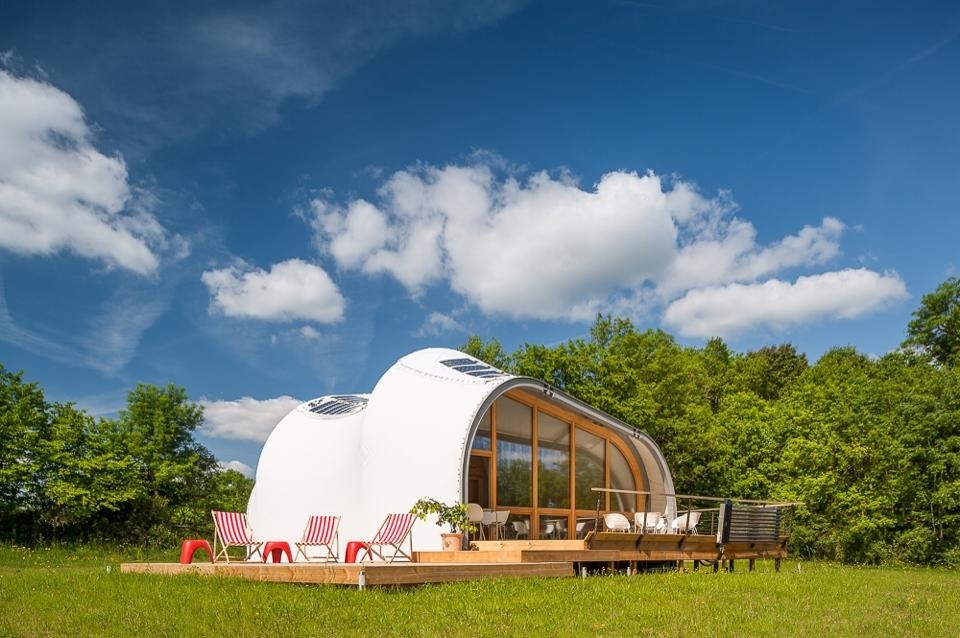 Cave-like house structure sitting in a field consisting of two conjoined oval pods. One wall is cut away to full-size windows. The house is surrounded by a wooden deck with red patio furniture.