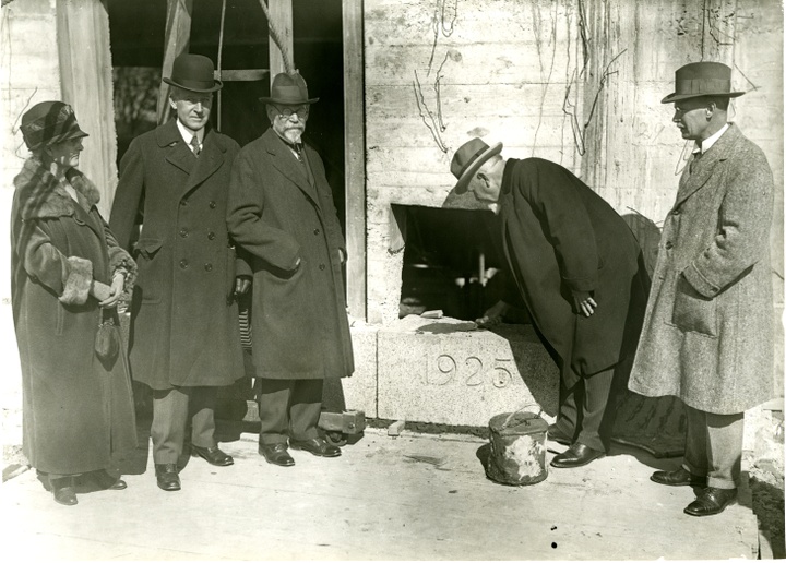 Black and white photograph of five people in dark coats and hats standing around a cornerstone block with "1925" engraved in it. One person is laying mortar the top.