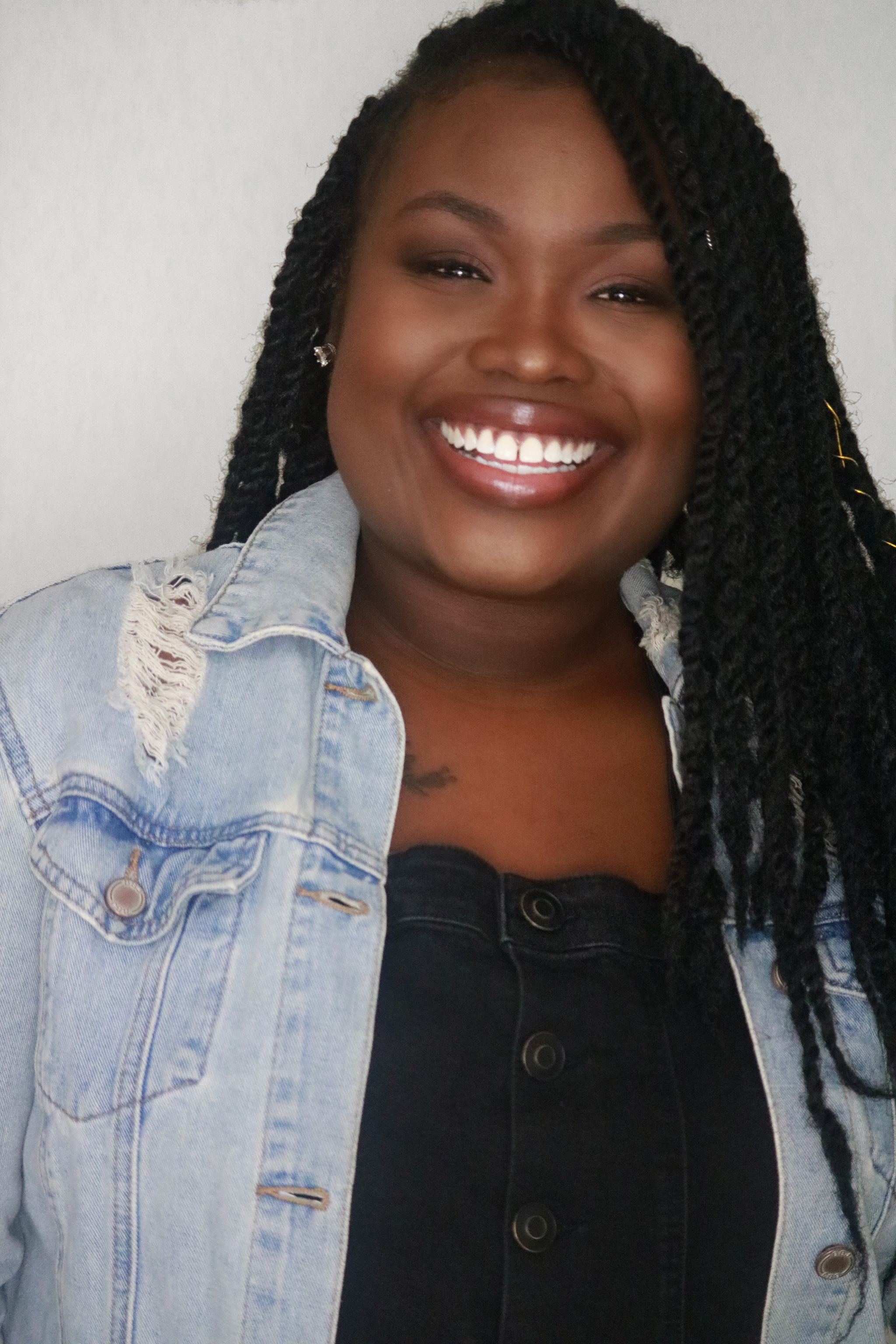 A Black woman with her hair in long braids smiles. She wears a light blue jean jacket and a black shirt underneath it.