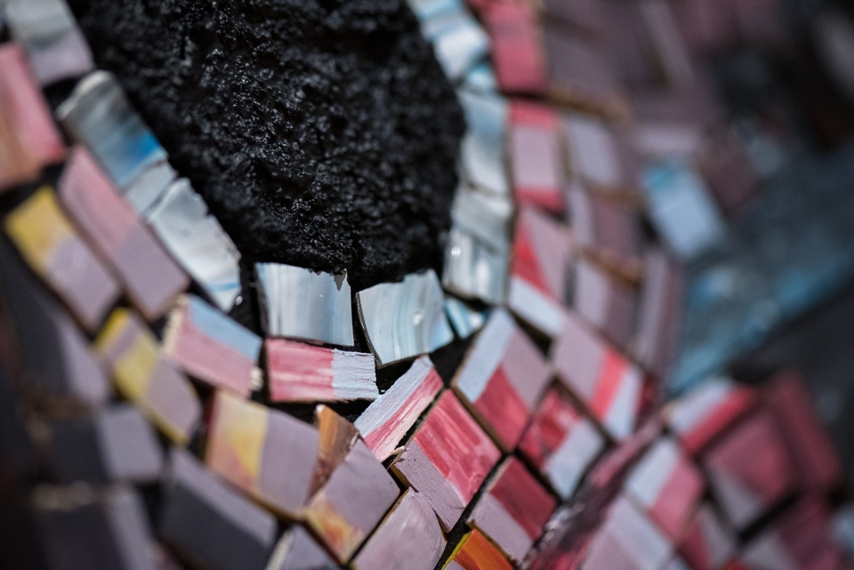 Detail shot of a mosaic arrangement of pink and blue shaded wooden chips embedded in a coarse, black substance with the texture of fresh asphalt.