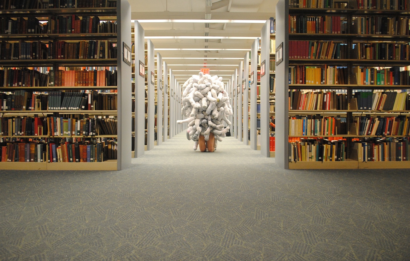 Person wearing a costume of white plush tubes covering their entire torso kneels on the floor between the stacks in a library.