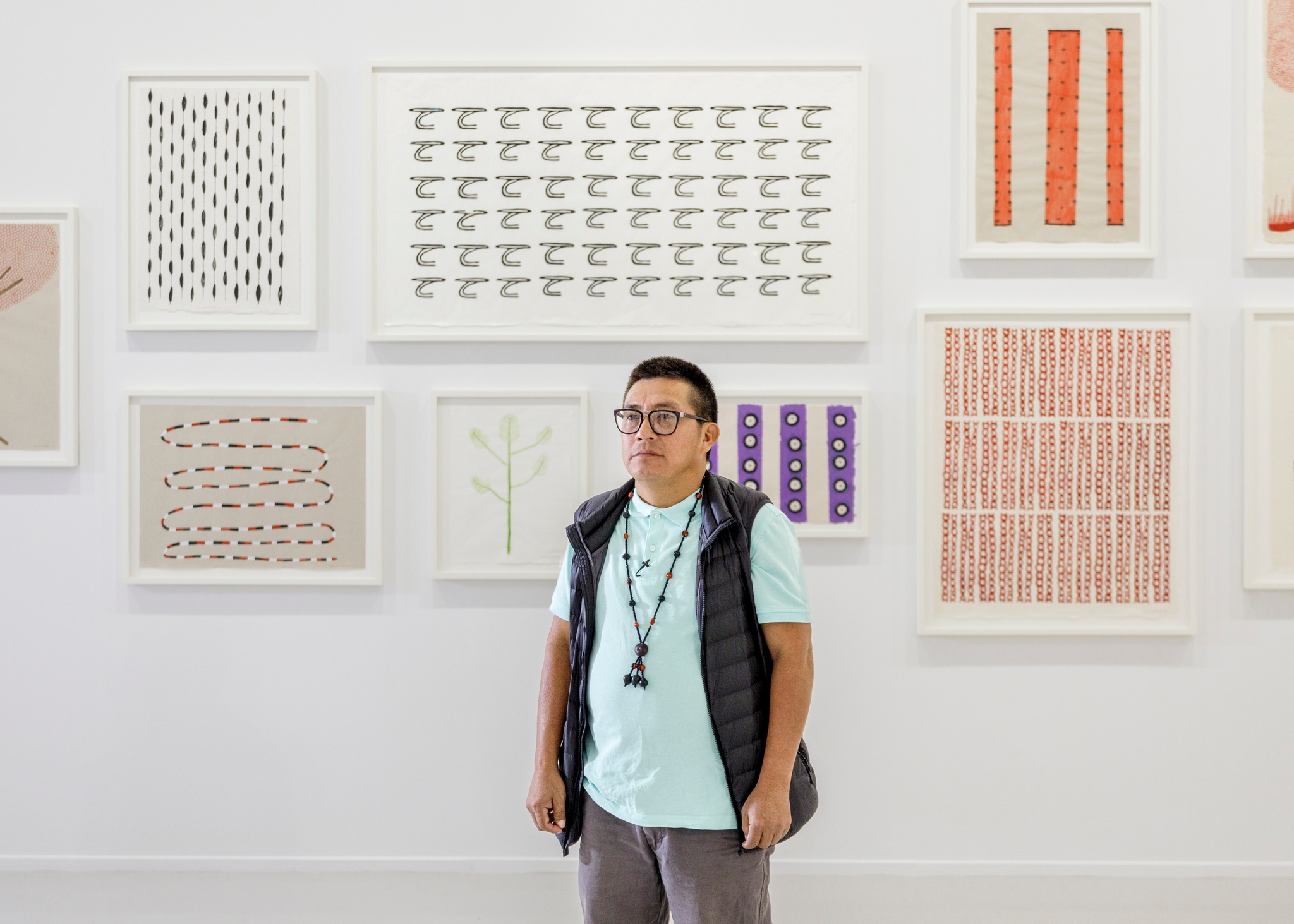 A portrait of Sheroanawe Hakihiiwe. An Indigenous Yanomami man, Sheroanawe poses in front of a gallery wall with numerous of his framed drawings. He wears a black puffer vest, a teal t-shirt, and a long beaded necklace that hags down nearly to his waist. He has short cropped dark hair and wears round-frame glasses. 