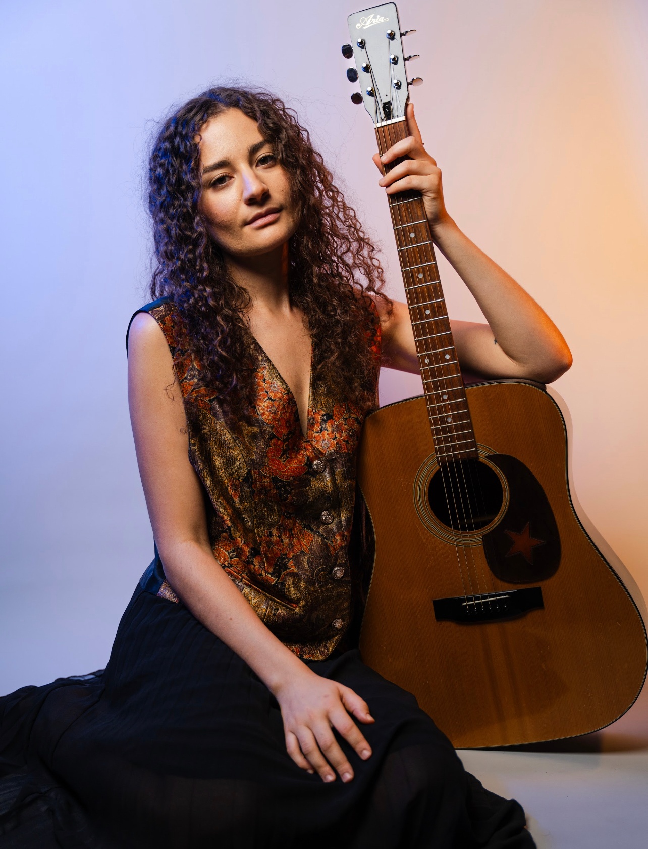 A white woman with curly brown hair falling below her shoulders looks at us contentendly while holding a guitar up at her side. She wears a silk vest with a shimmering deep brown and orange pattern. Behind her a wall is lit in purple and orange light.