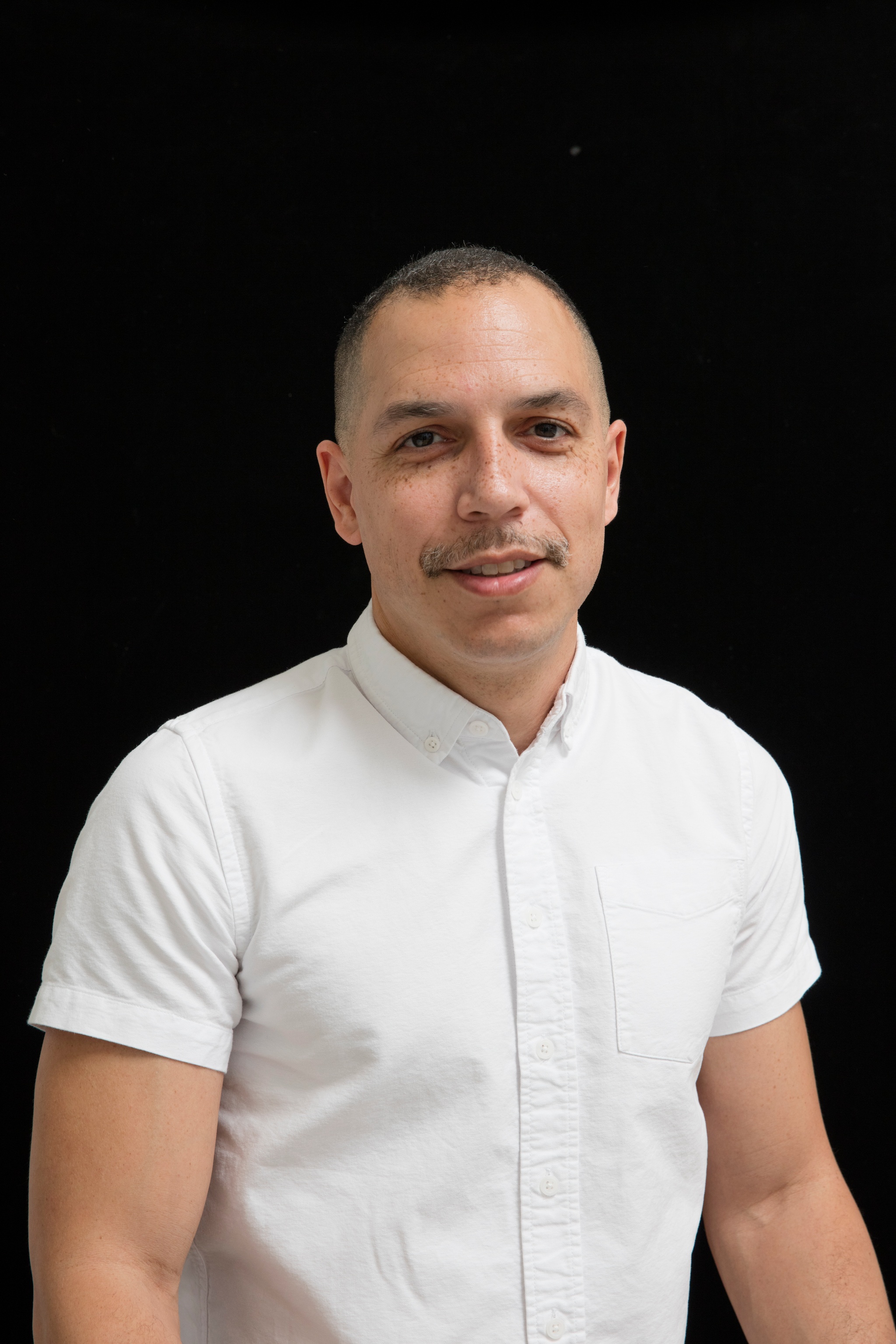 A headshot of Malik Gaines. He poses against a black background and wears a white short sleeved button down shirt buttoned to the neck. He smiles with his mouth slightly open. He has a faint mustache. 