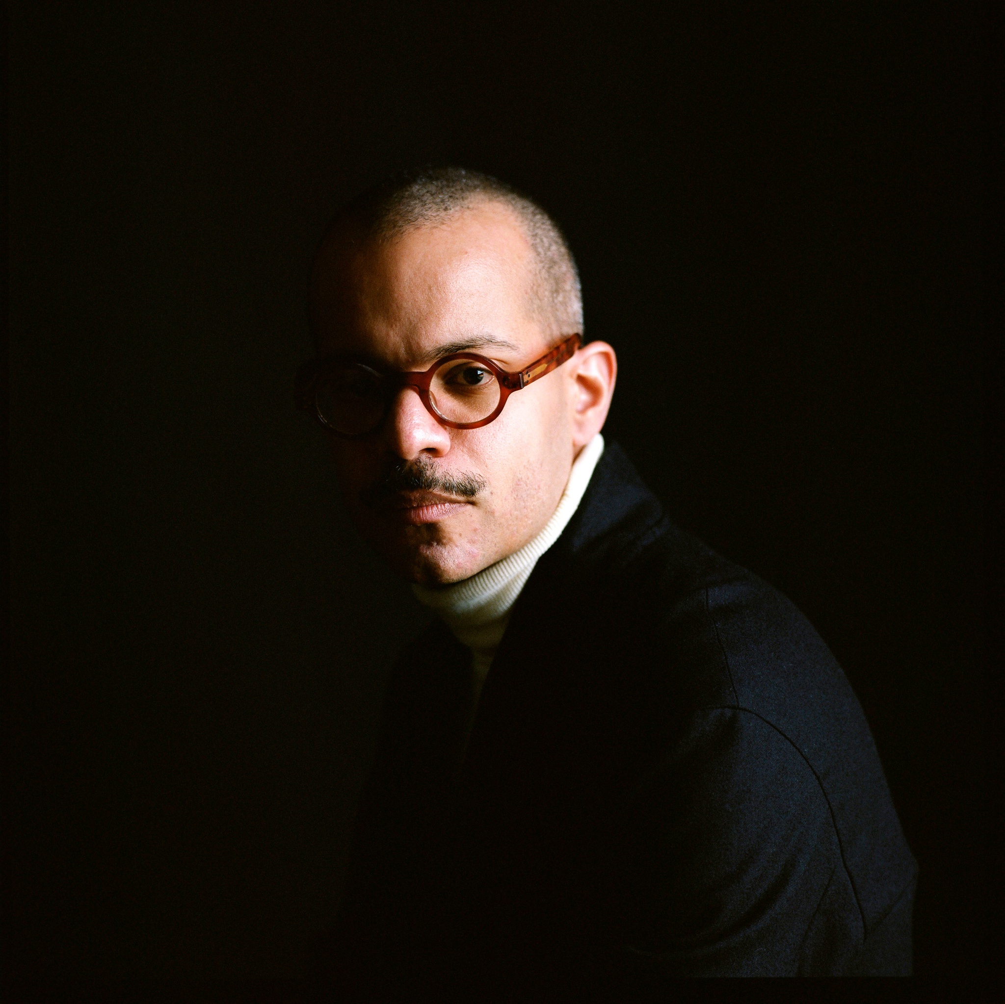A portrait of costume designer Carlos Soto. He sits in half shadow, with the left side of his face obscured. He wears round-rimmed glasses and a turtleneck and has a head shaved close to the scalp. 