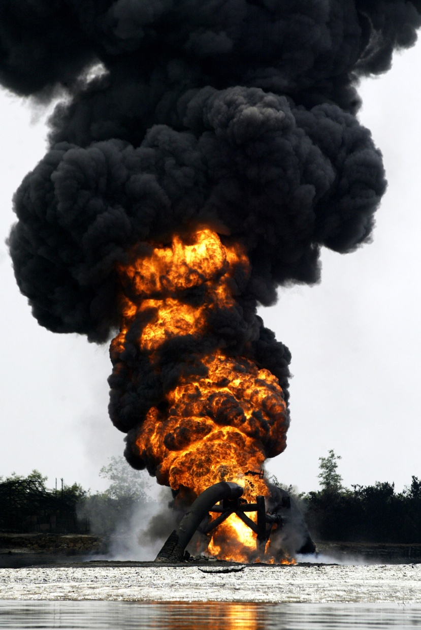 Photograph of a large plomb of thick black smoke coming from a burning structure that appears to be sitting on top of water.
