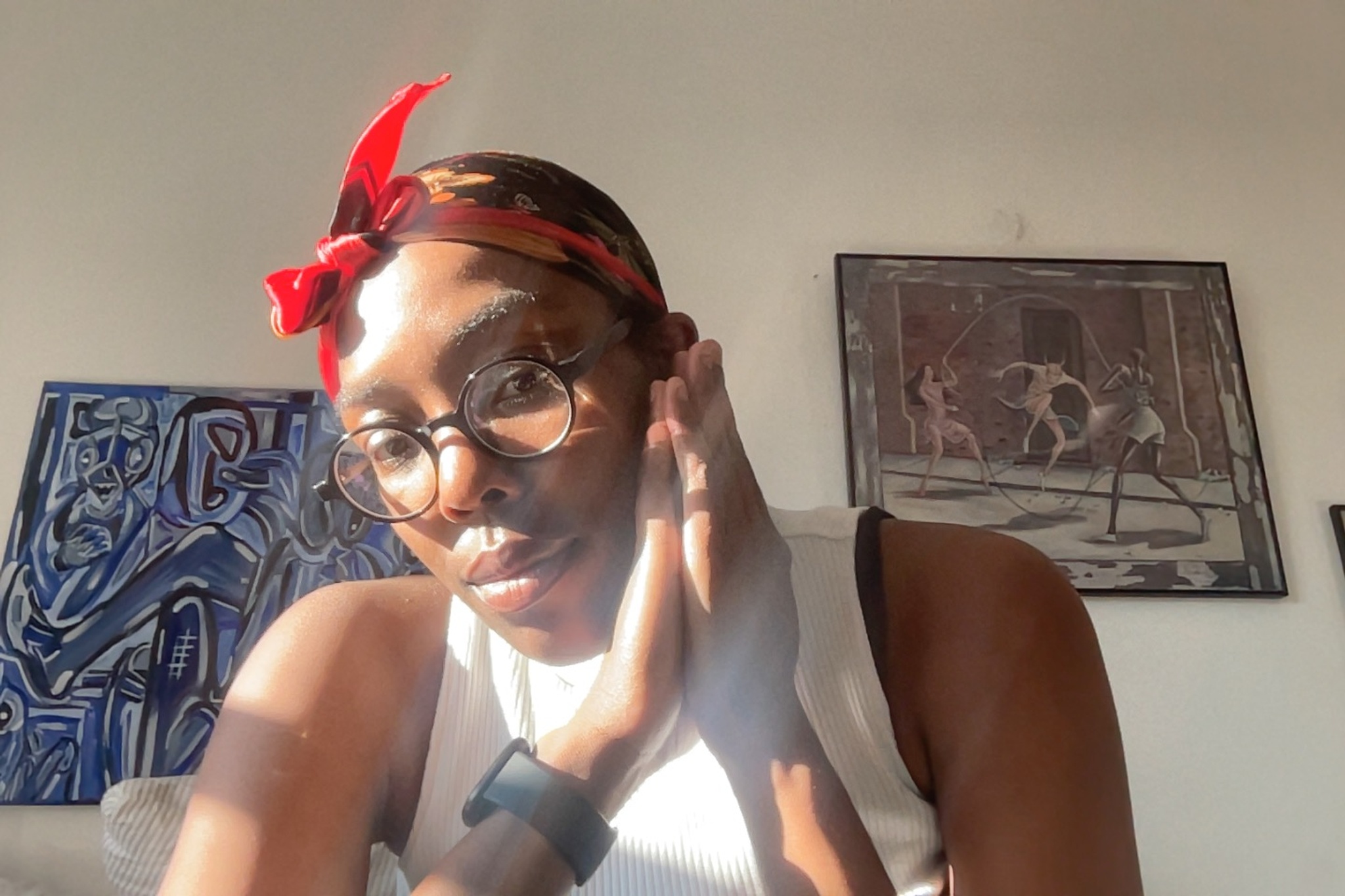 This is a headshot of Joselia Rebekah Hughes, who is an Afro-Caribbean-descended Black woman. She is posing in bright sunlight from a window in front of a wall with art hanging on it. She leans slightly forward, holding her hands flat together beneath her chin, with her gaze looking down at us. She wears glasses, a white sleeveless t-shirt, and a red scarf that is tied above her forehead. 