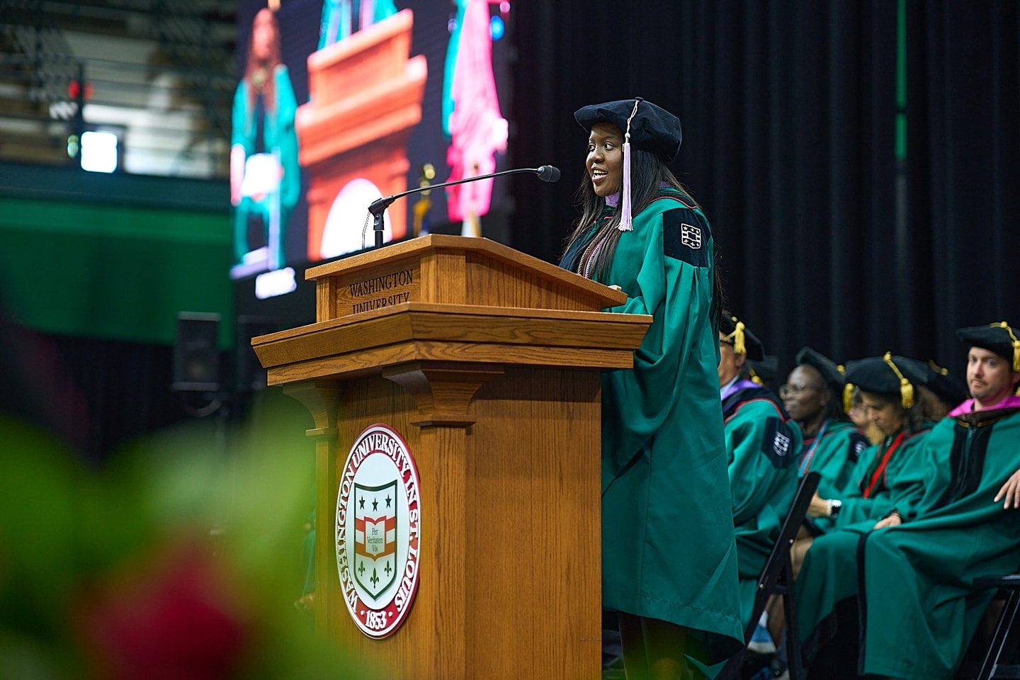 A woman wearing regalia delivers an address from behind a lectern.