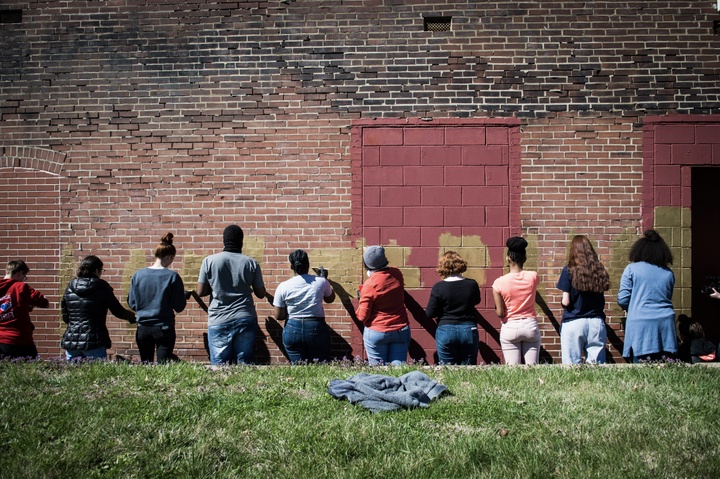 A group of 10 people painting an old, red-brick wall gold.