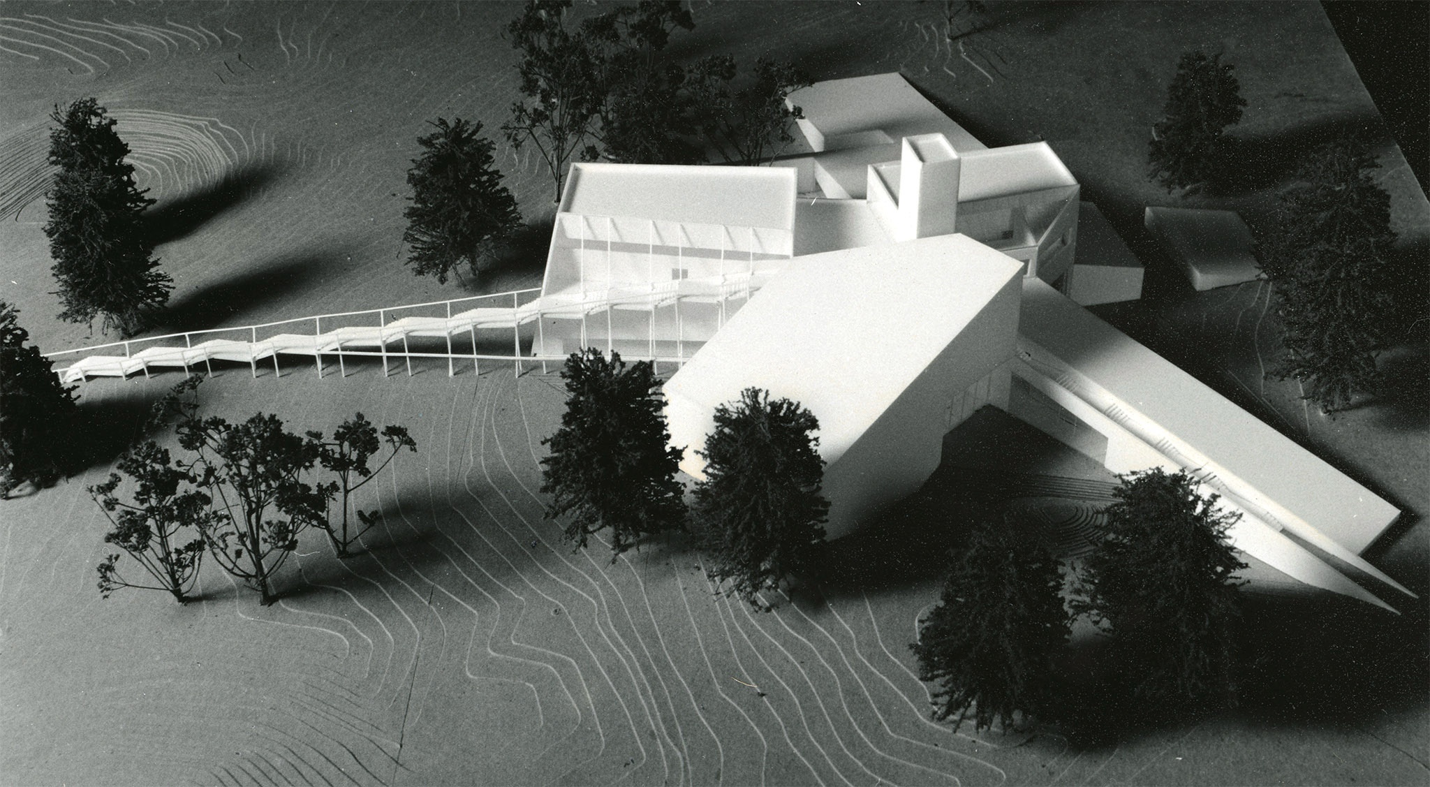 A black and white, side-aerial photograph of a small, white, model of an angular building surrounded by fake trees.