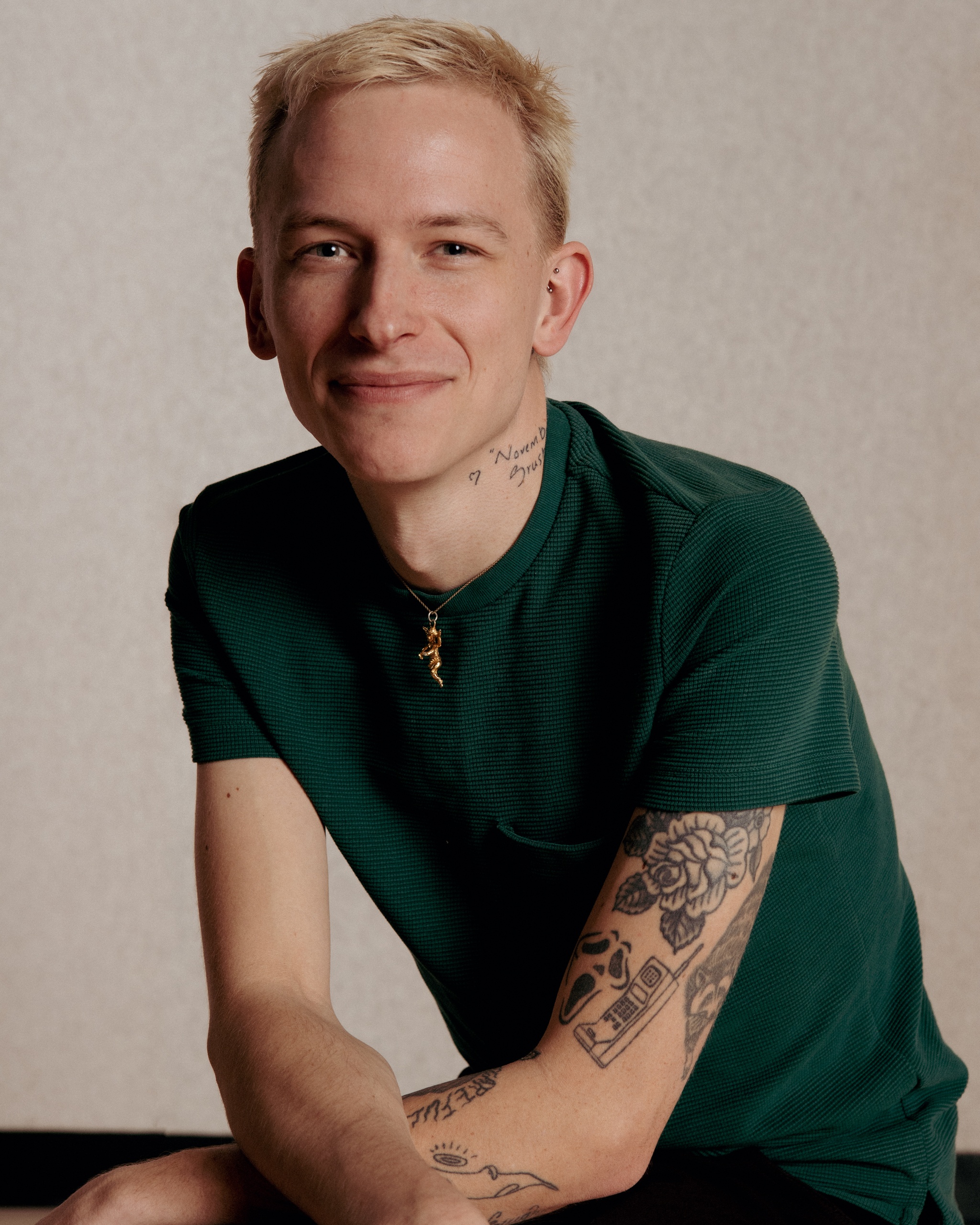 A portrait of Jake Brush, a white person posing with arms loosely crossed across his knee. He has short bleached blond hair and smiles with his eyes without opening his mouth. He wears a dark green tshirt and has tattoos down his left arm. Photo by Dana Golan