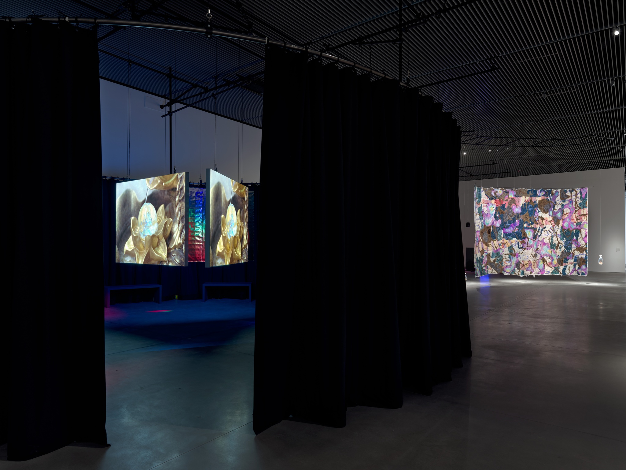 Two artworks in a gallery. On the left, a triangular formation of three video screens hangs inside a circular area defined by heavy dark drapes. On the right in the background hangs a multicolored abstract felt tapestry.