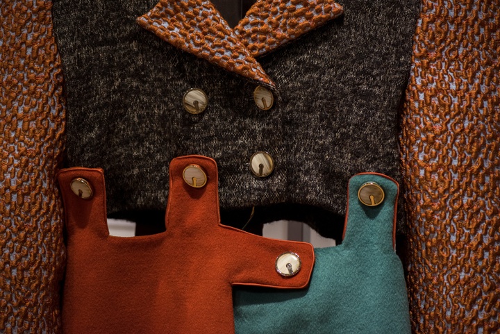 Detail view of a double-breasted garment constructed of several pieces of heavy, textured fabrics in rust red, teal, and brown, held together by fabric tabs and buttons.