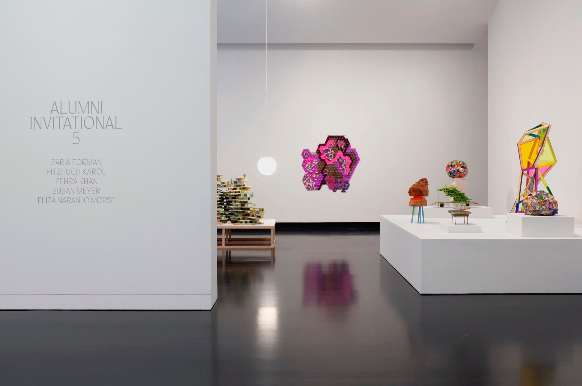 An interior of a museum gallery with one wall showing the title Alumni Invitational 5, with a number of colorful sculptures sitting on various plinths, and hung on the wall.