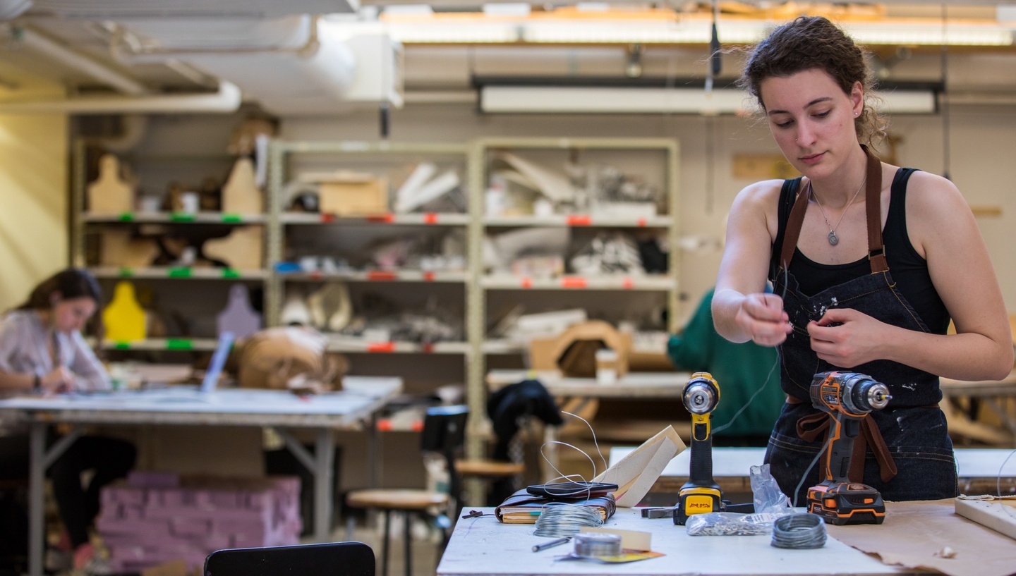 Person wearing an apron works with a piece of wire and a drill in a shop space.