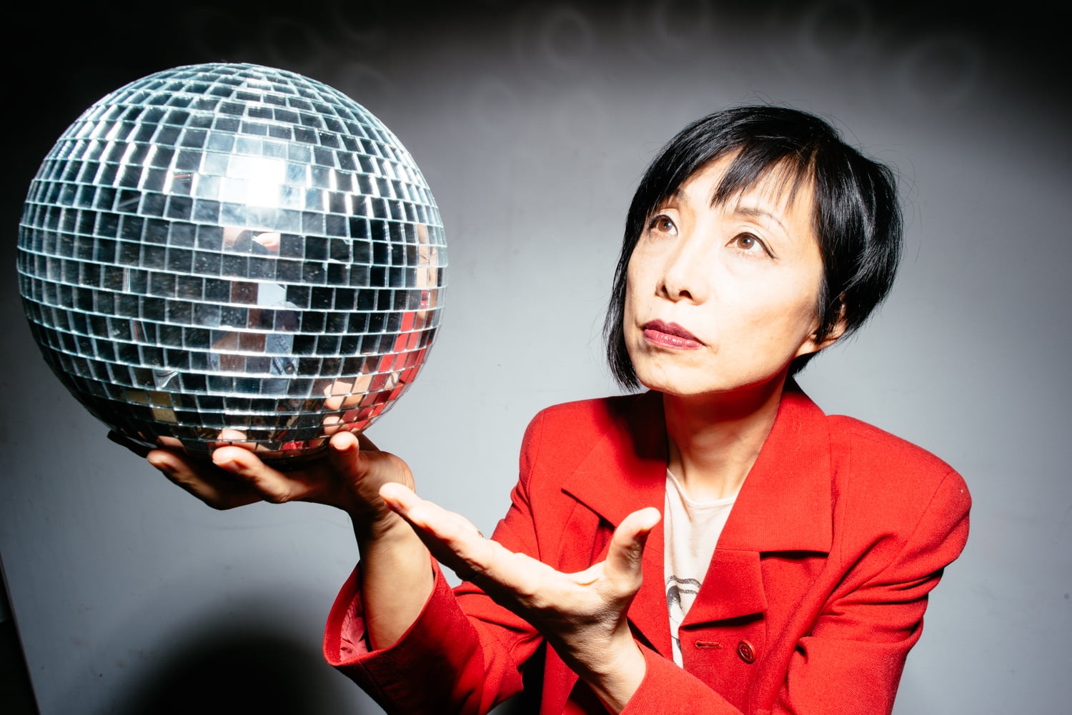 A Japanese American woman with short black hair reaches out to hold a disco ball in one hand. She wears a bright red jacket and looks toward the disco ball.