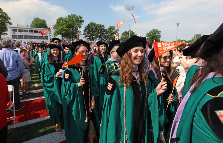 Students in green graduation robes wave orange flags reading "Sam Fox School" as they process down a football field flanked by clapping attendees.