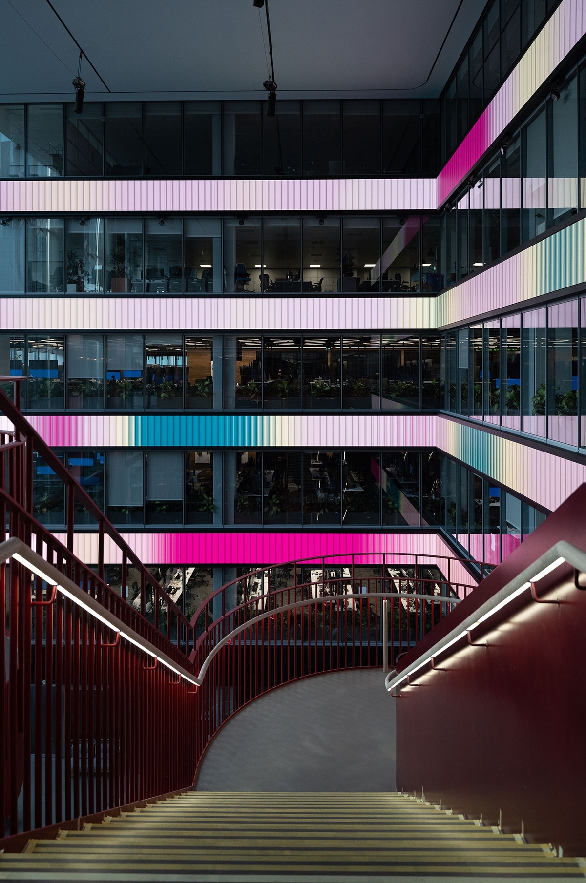 View from top of stairwell, overlooking installation in the atrium of four long LED screens between four floors of the building, with pink, green and blue gradients