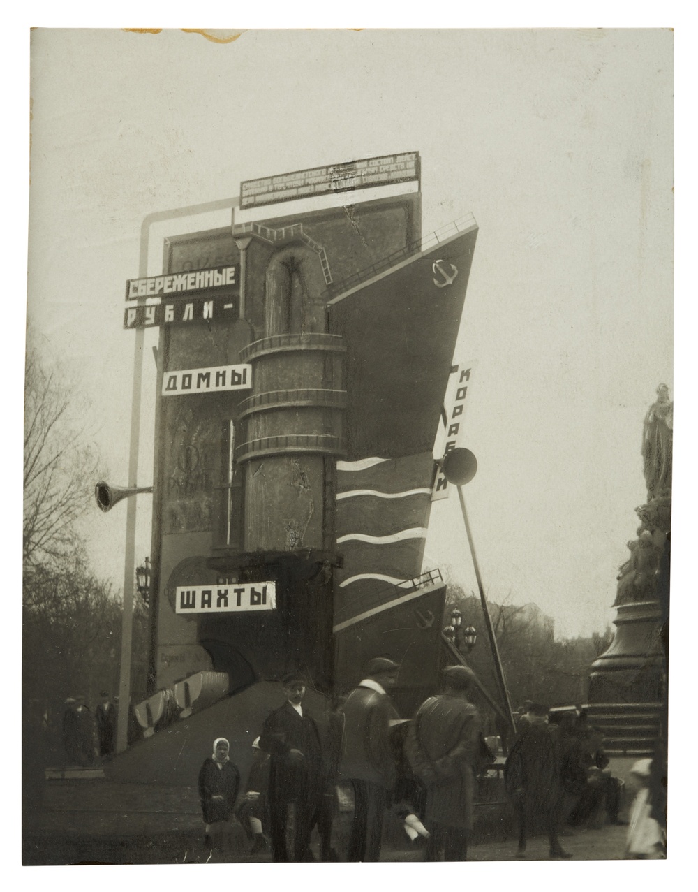 Black and white photograph of an abstract building structure with Russian signage, possibly in a park with people walking around it.