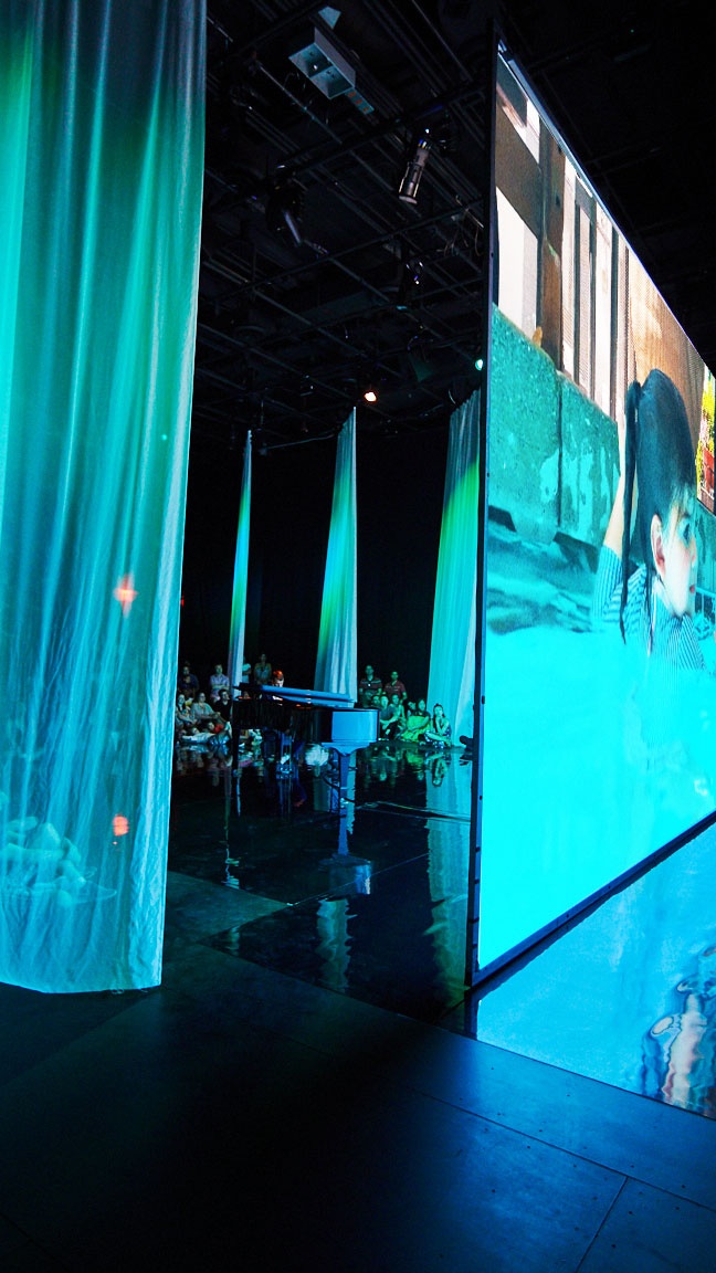 Audience viewing film on large screen in blue-lit space.