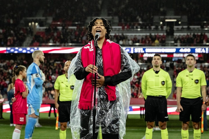 A person sings into a microphone in a soccer stadium while players stand behind her with their arms behind their backs.