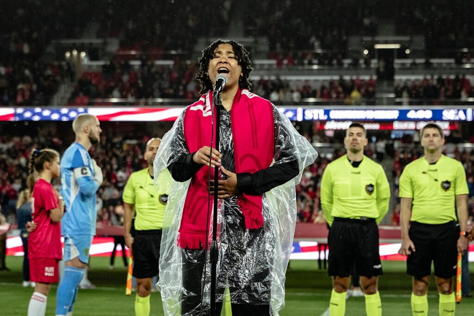 A person sings into a microphone in a soccer stadium while players stand behind her with their arms behind their backs.